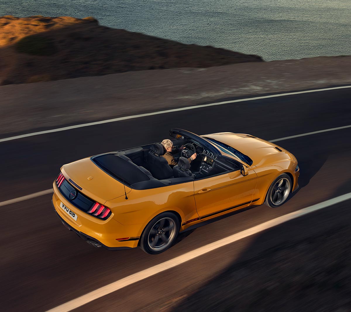 Ford Mustang California arancione. Vista posteriore a tre quarti, in marcia su una strada.