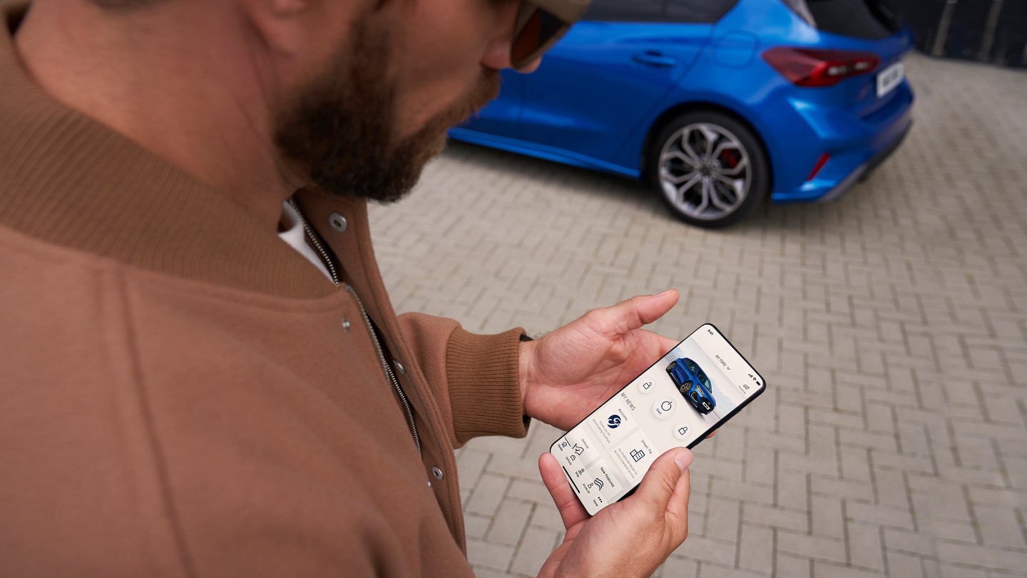 App FordPass. Un uomo guarda il proprio smartphone. Sullo sfondo una Ford Focus blu.