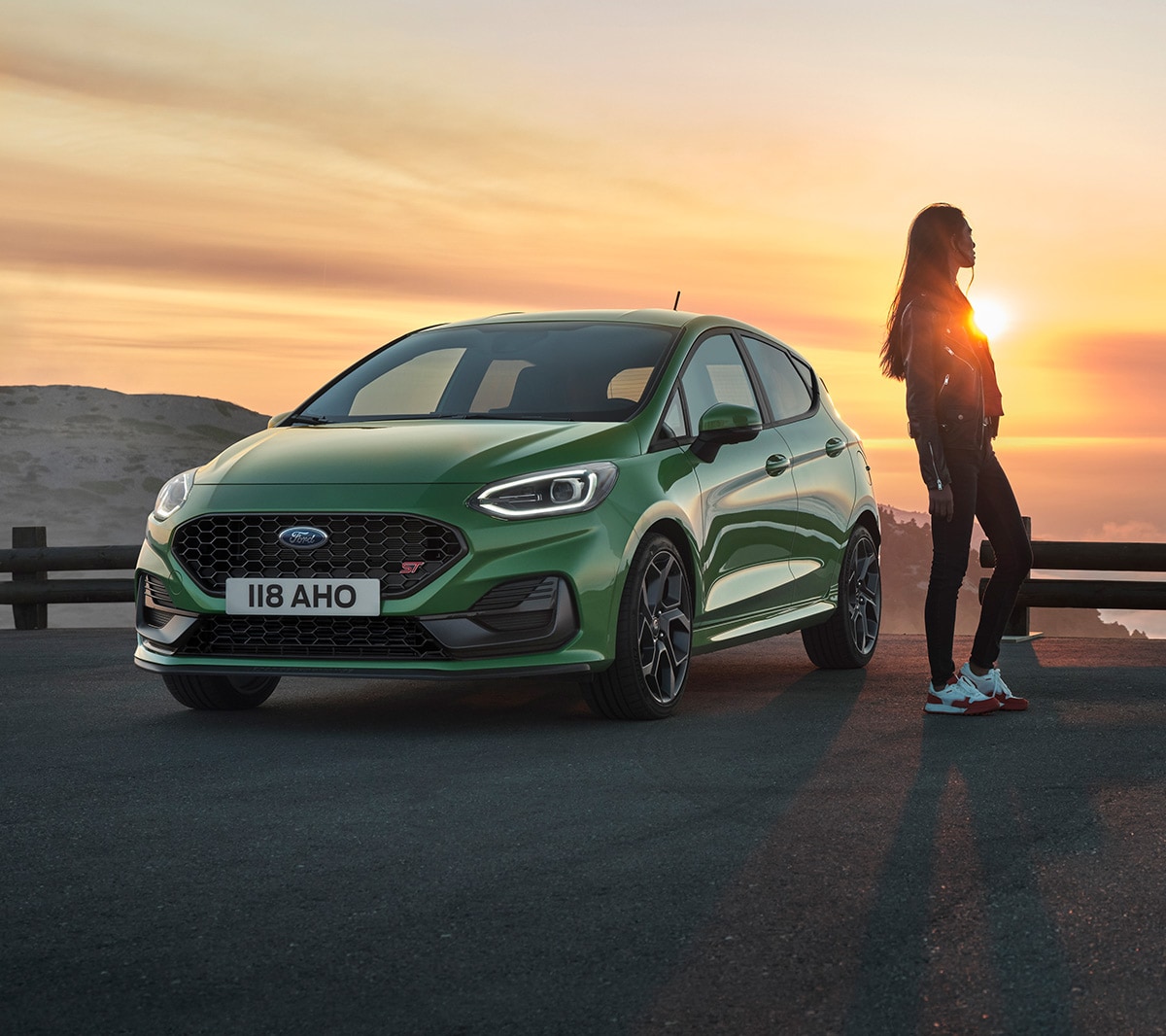 Ford Fiesta ST verde. Vista frontale a tre quarti, una donna ammira il tramonto.