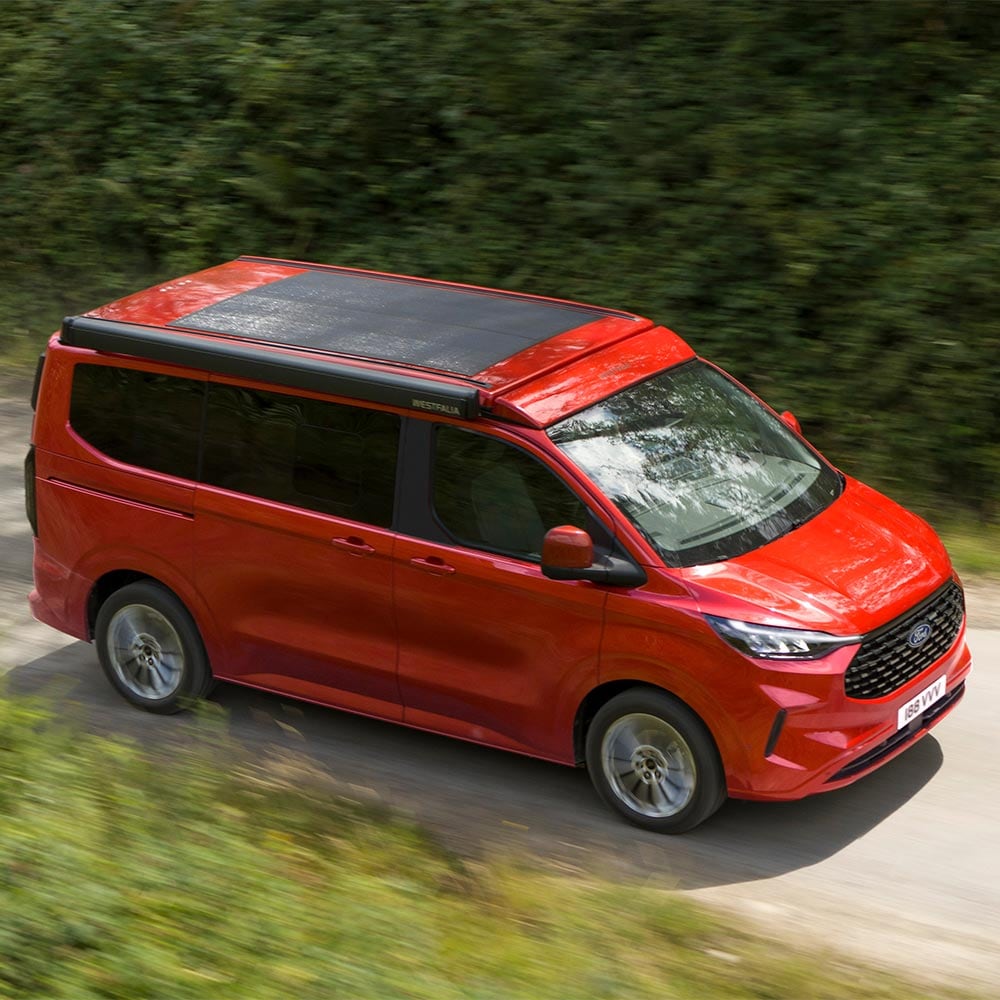 Ford Nugget rosso con pannello solare sul tetto percorre la strada.
