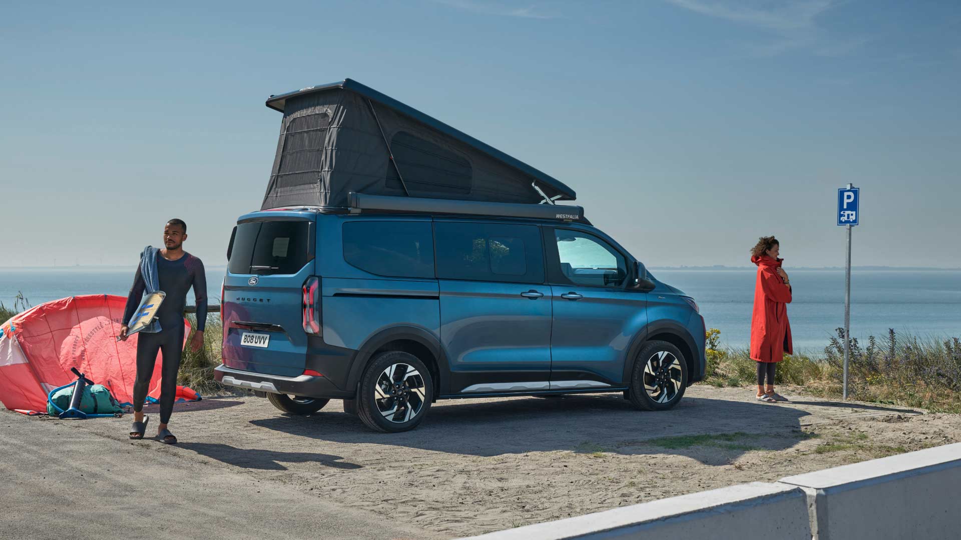 Un Ford Transit Custom blu con tetto a soffietto è parcheggiato al mare. Due persone e una tavola da surf sono vicine.