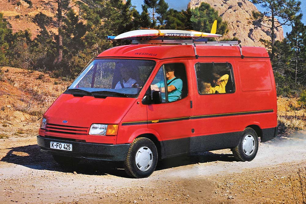 Un Ford Nugget del 1993 con portapacchi e tavola da surf percorre una strada polverosa tra le montagne.