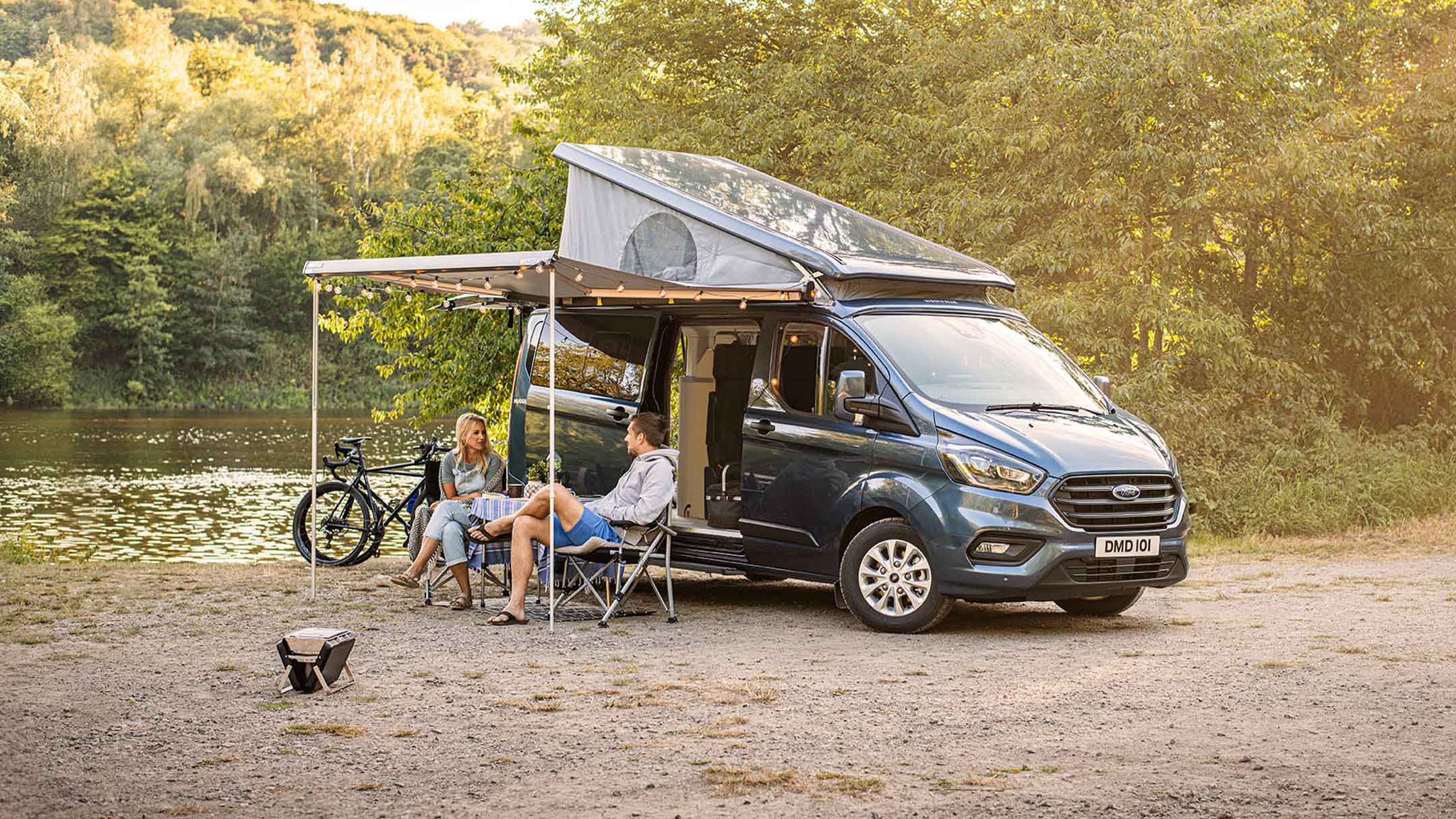 Ford Transit Custom Nugget blu con tetto apribile in vista laterale con persone sedute sotto la tettoia, parcheggiato in riva a un lago