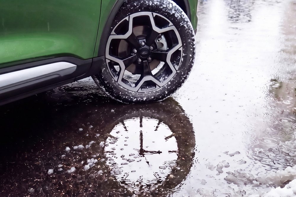 La ruota anteriore di una Ford Kuga verde si trova accanto a una pozzanghera, nella cui superficie dell’acqua si riflette un quadrante di orologio.