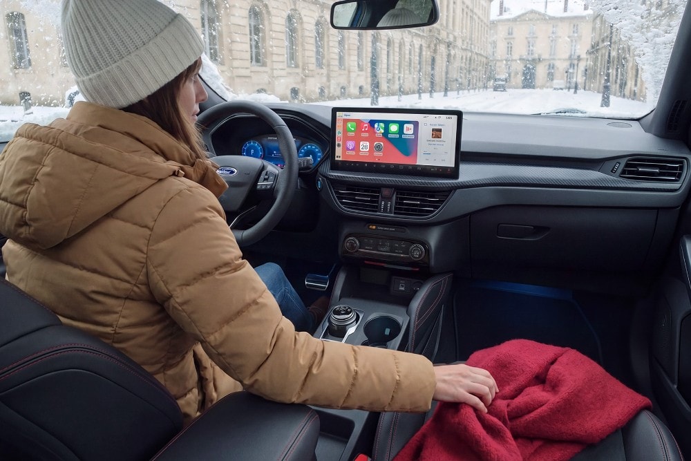 Una donna con un cappotto invernale e un berretto è seduta in una Ford in una giornata nevosa, tenendo una coperta rossa.