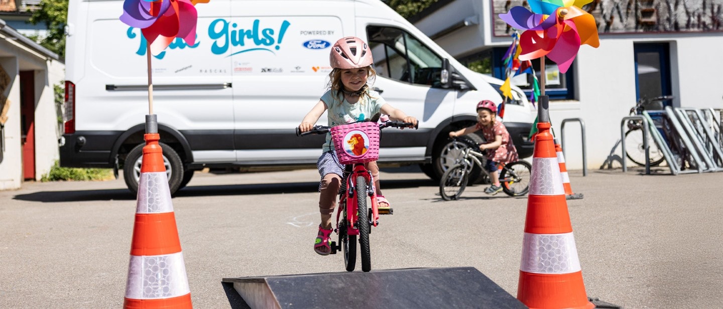 Una bambina pedala su una bicicletta in un percorso a ostacoli. Sullo sfondo una Ford E-Transit.