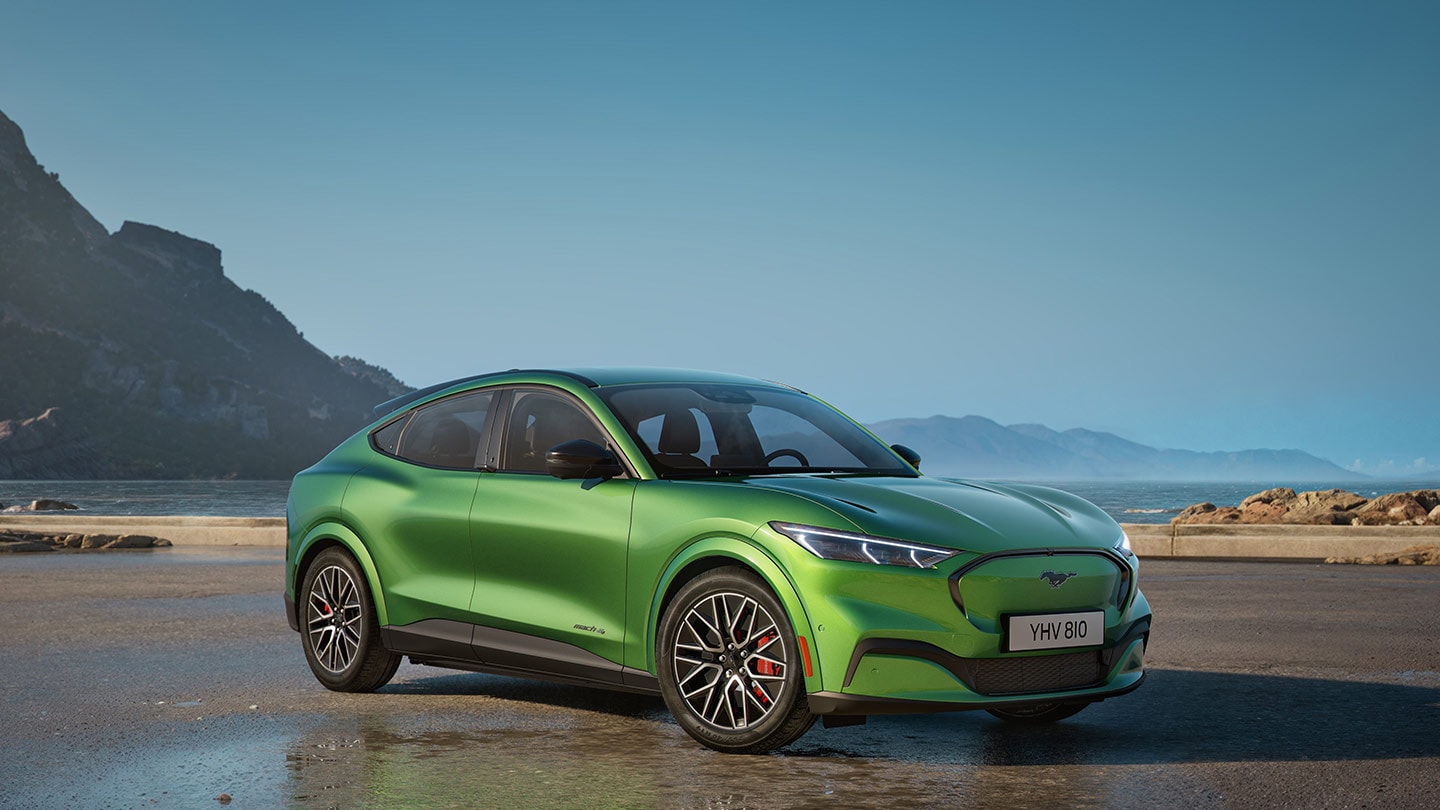 Vista laterale anteriore di una Ford Mustang Mach-E verde parcheggiata vicino a uno specchio d'acqua, con montagne sullo sfondo.