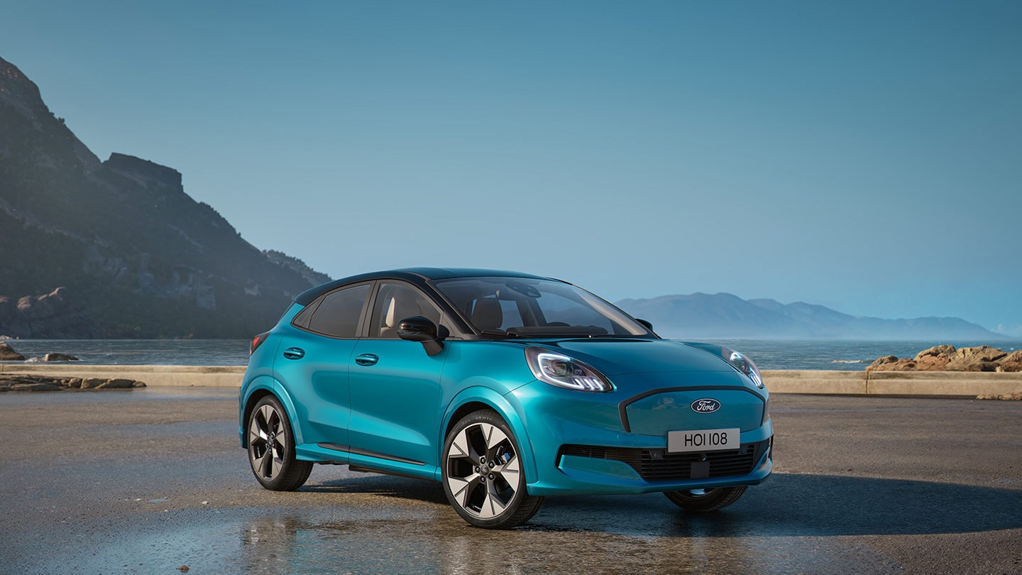 Vista frontale di una Ford Puma Gen-E blu parcheggiata vicino a uno specchio d'acqua, con montagne sullo sfondo.