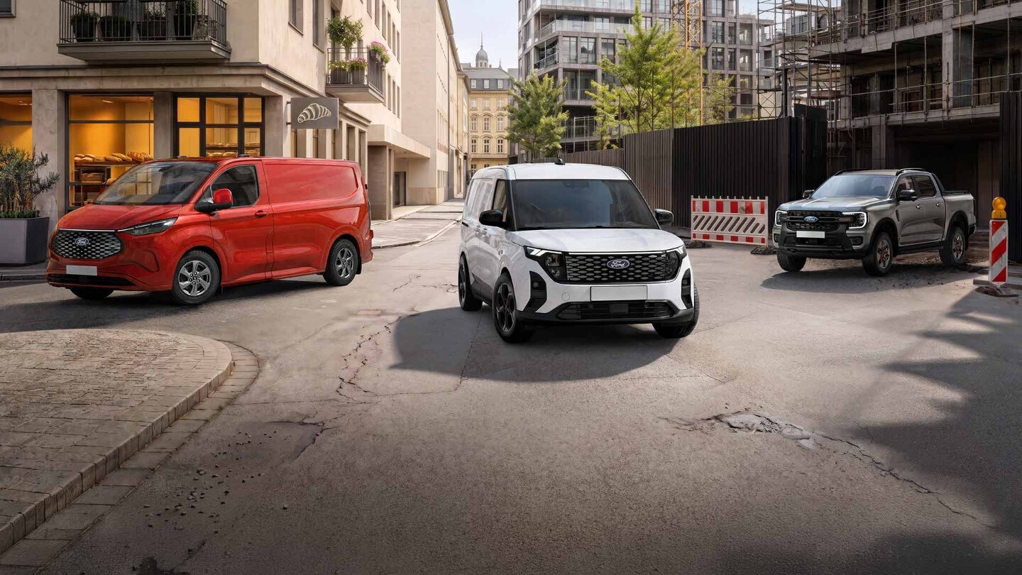 Tre veicoli Ford, un Transit Custom rosso, un E-Transit Custom bianco e un Ranger grigio, sono parcheggiati su una strada urbana.
