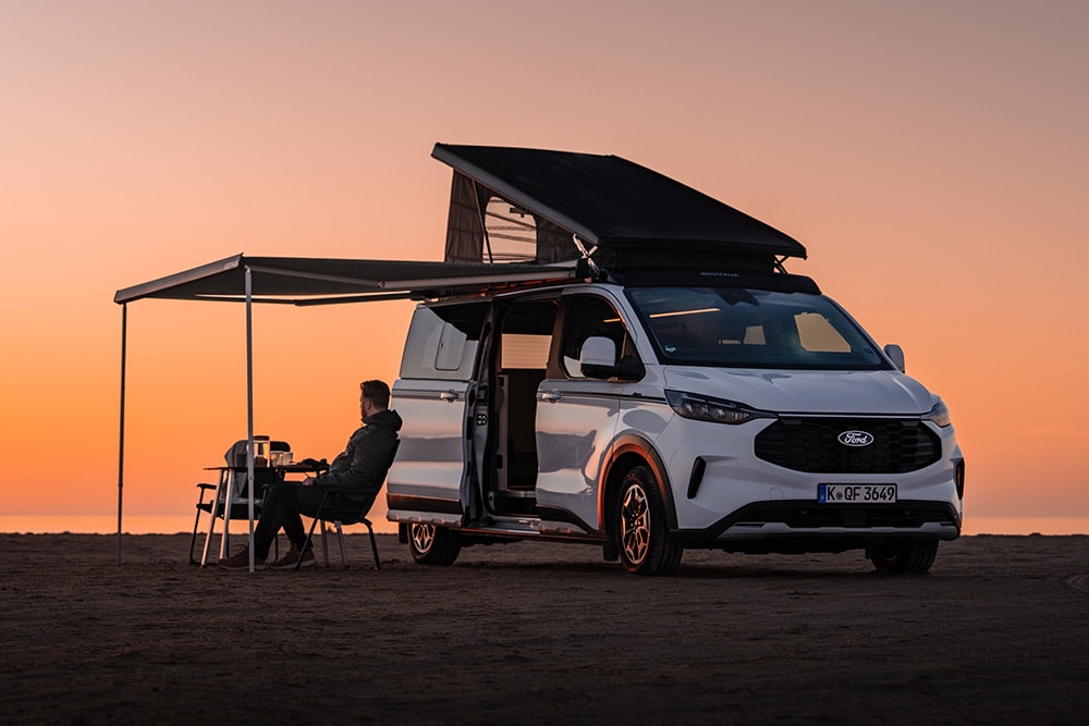 Un Ford Nugget in edizione anniversario con tetto e tenda aperti in un paesaggio sconfinato al crepuscolo. Una persona siede su una sedia da campeggio sotto la tenda.