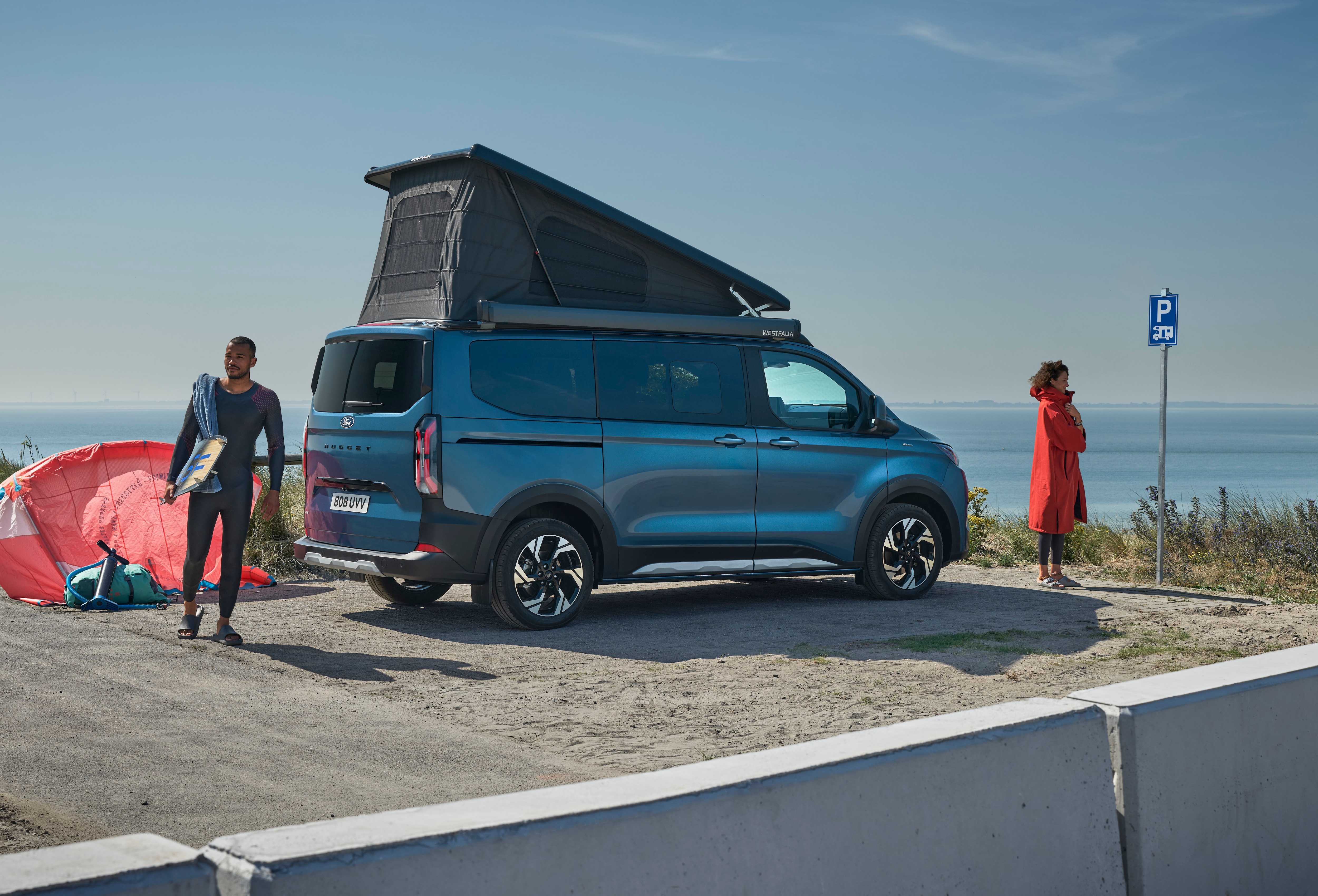 Un Ford Nugget blu scuro con tetto aperto è parcheggiato al mare mentre due persone preparano l’attrezzatura per il kitesurf.
