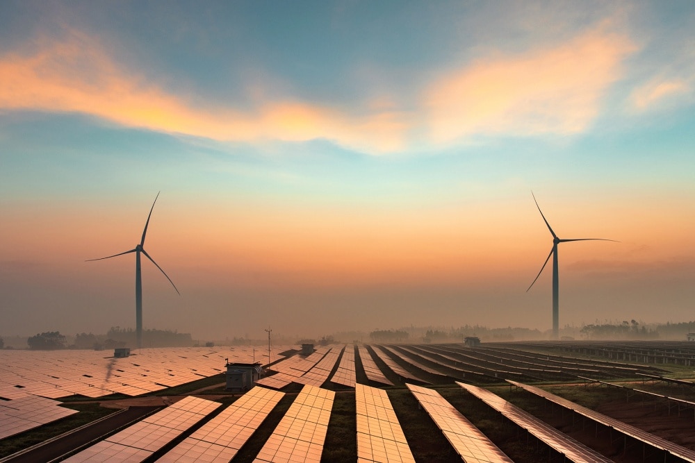Un campo pieno di pannelli fotovoltaici con due pale eoliche all’orizzonte