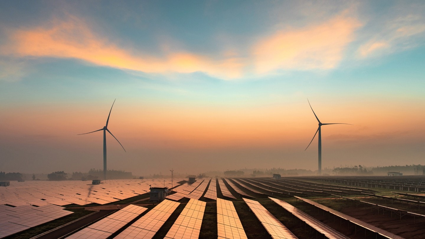 Un campo pieno di pannelli fotovoltaici con due pale eoliche all’orizzonte