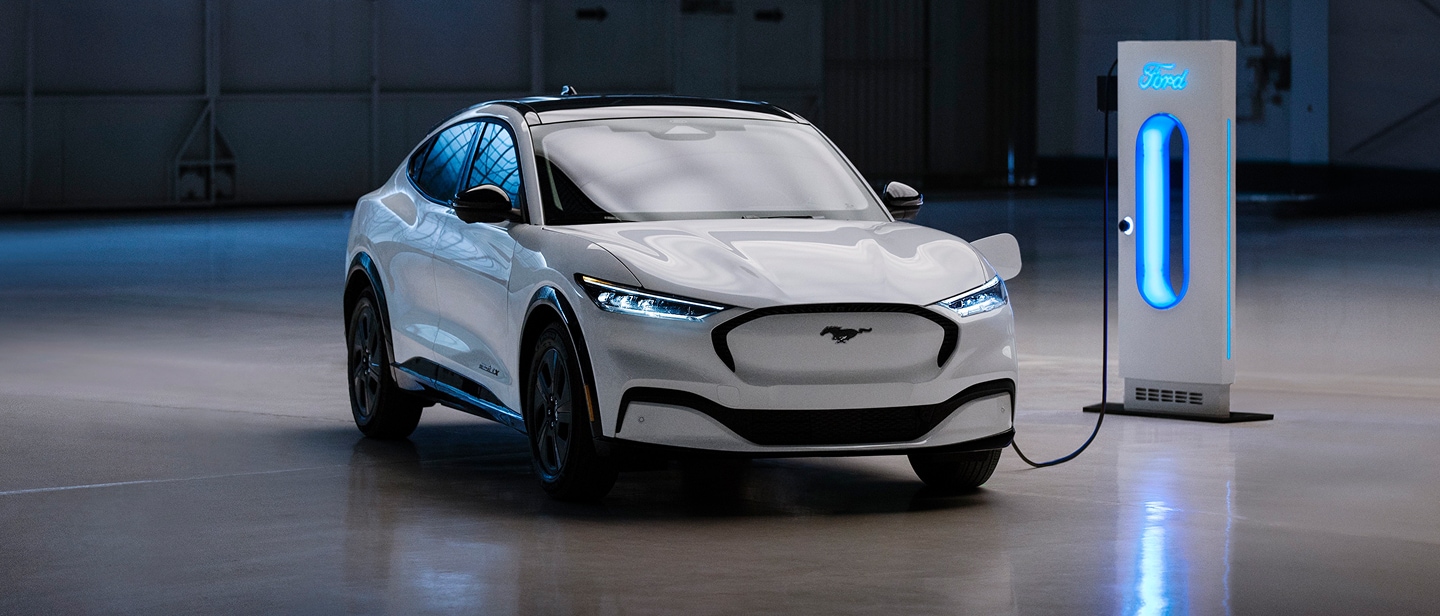 Una Ford Mustang Mach-E bianca è parcheggiata in un garage ed è collegata a una stazione di ricarica illuminata di blu.