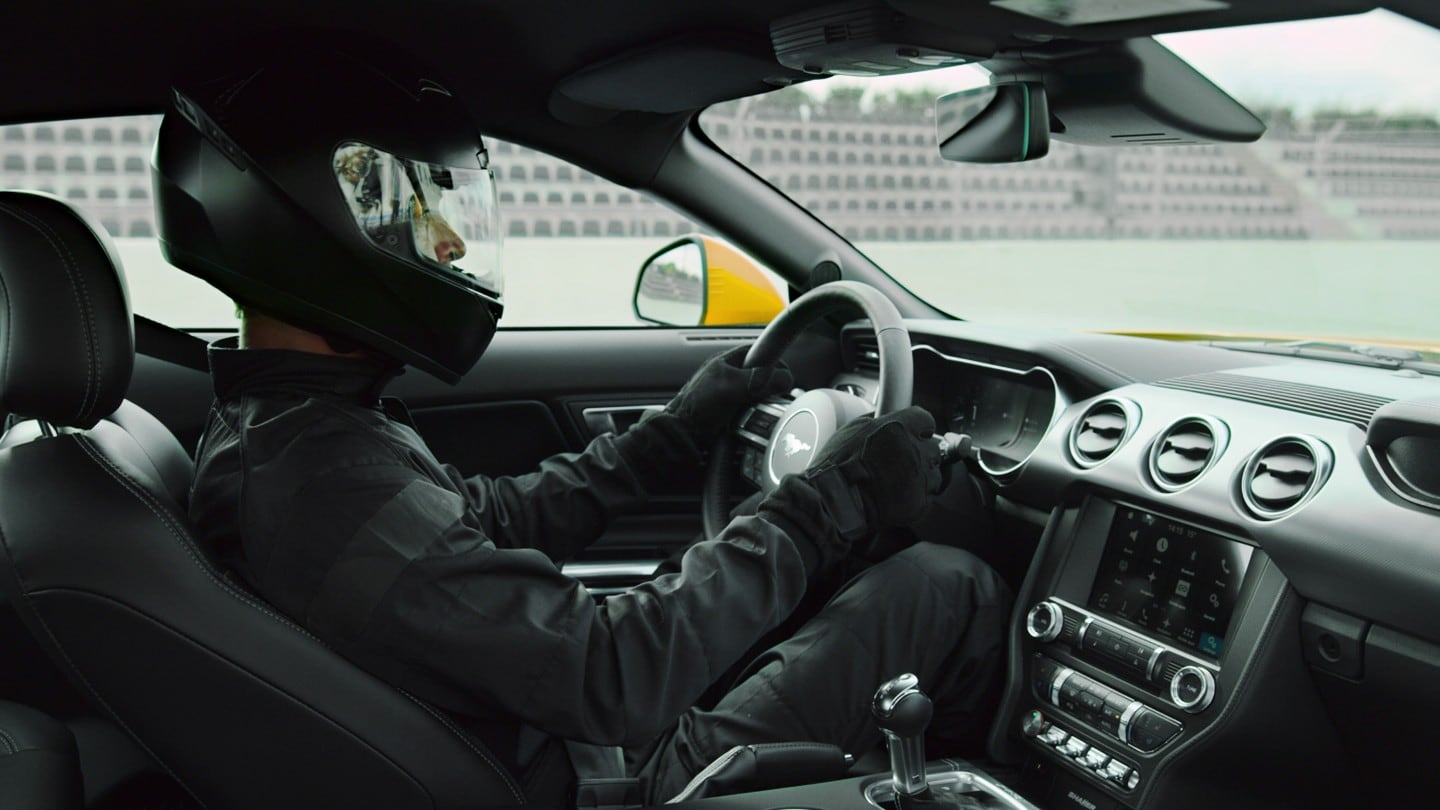 Ford Mustang. Vista degli interni dal sedile del passeggero, con un pilota sportivo al volante che indossa un casco nero