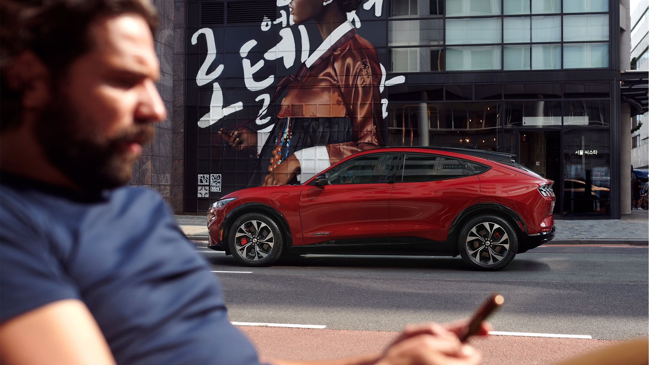 Ford Mustang Mach-E rossa. Vista laterale. In primo piano un uomo che guarda il proprio smartphone