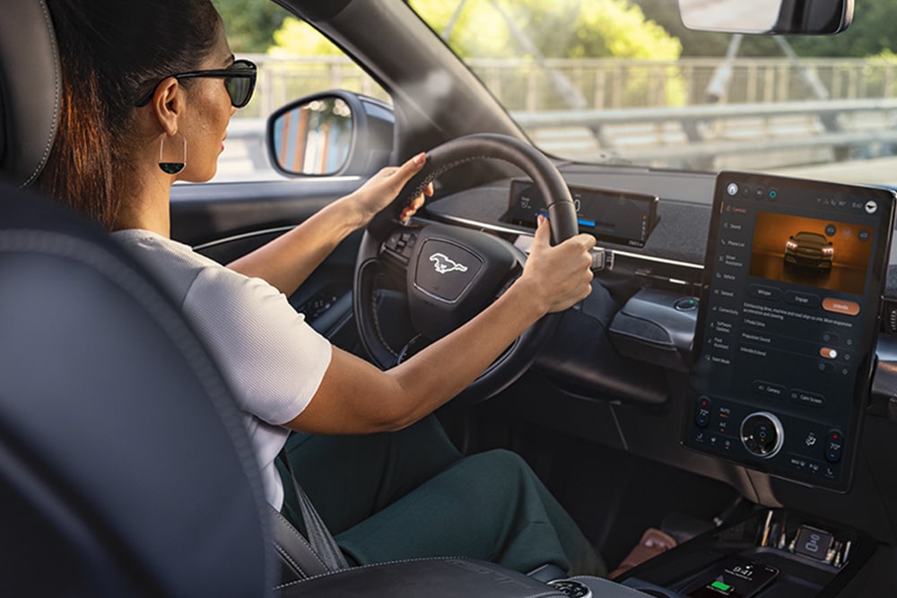 Una donna guida un Mustang Mach-E con le mani sul volante. Il touchscreen da 15,5" mostra i dati del veicolo.