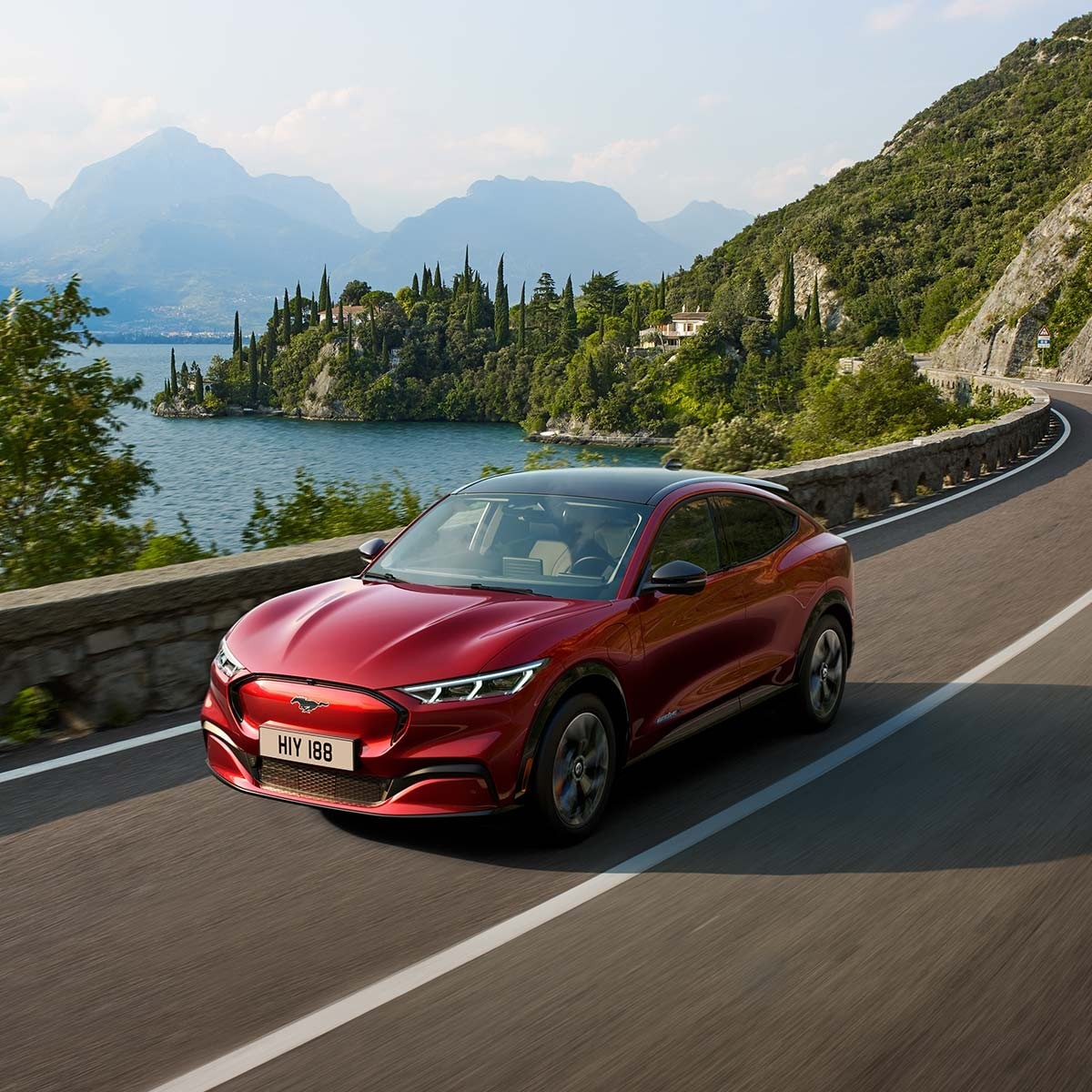 Ford Mustang Mach-E rossa. Vista a tre quarti, di passaggio su strada montana davanti a un lago
