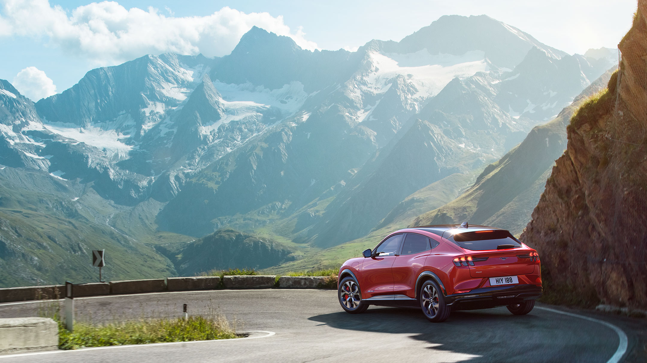 Ford Mustang Mach-E rossa. Vista posteriore a tre quarti. Ambientazione su una strada panoramica montana tortuosa