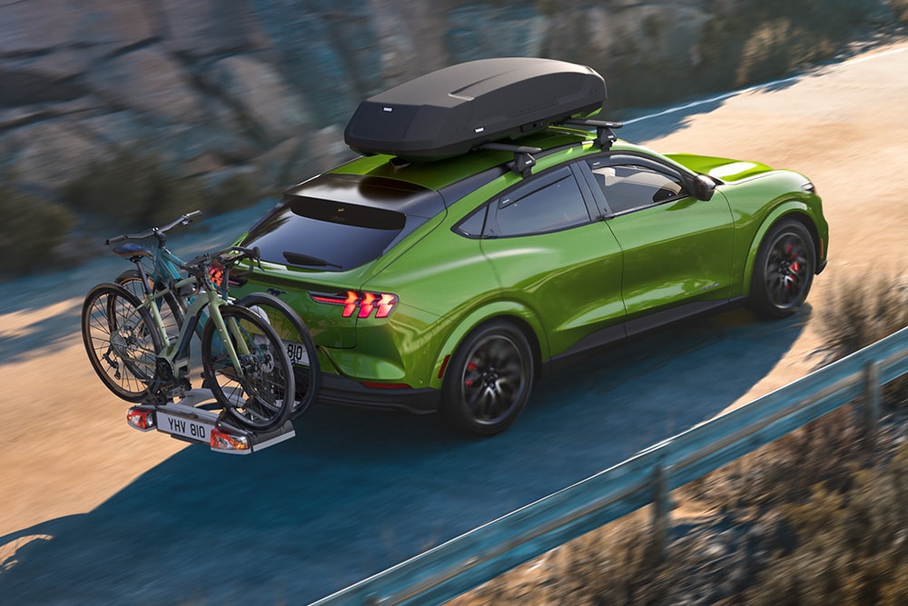 Ford Mustang Mach-E verde con portabiciclette e box da tetto su una strada tortuosa.