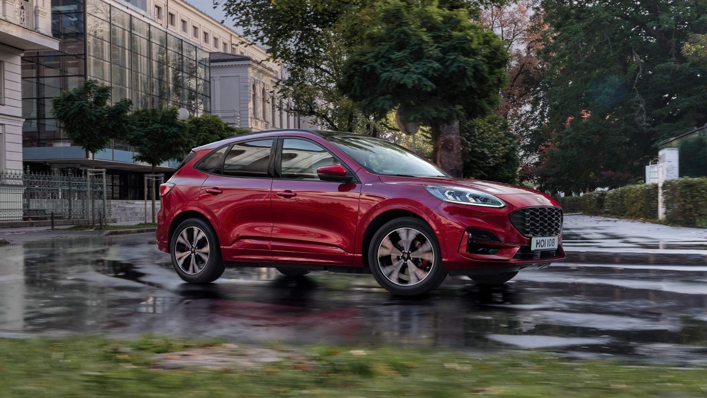 Ford Kuga rossa, vista frontale a tre quarti, in marcia in città con edifici moderni e alberi sullo sfondo