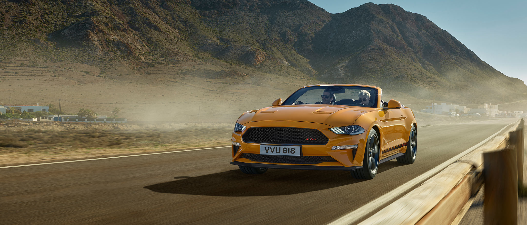 Ford Mustang California arancione. Vista frontale a tre quarti, in marcia in mezzo alla natura con montagne sullo sfondo.