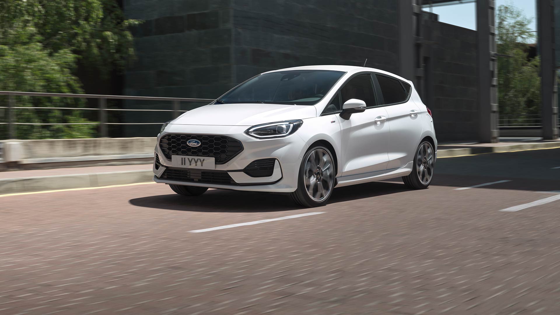 Ford Fiesta bianca che percorre una strada cittadina, con vegetazione ed edifici sullo sfondo.