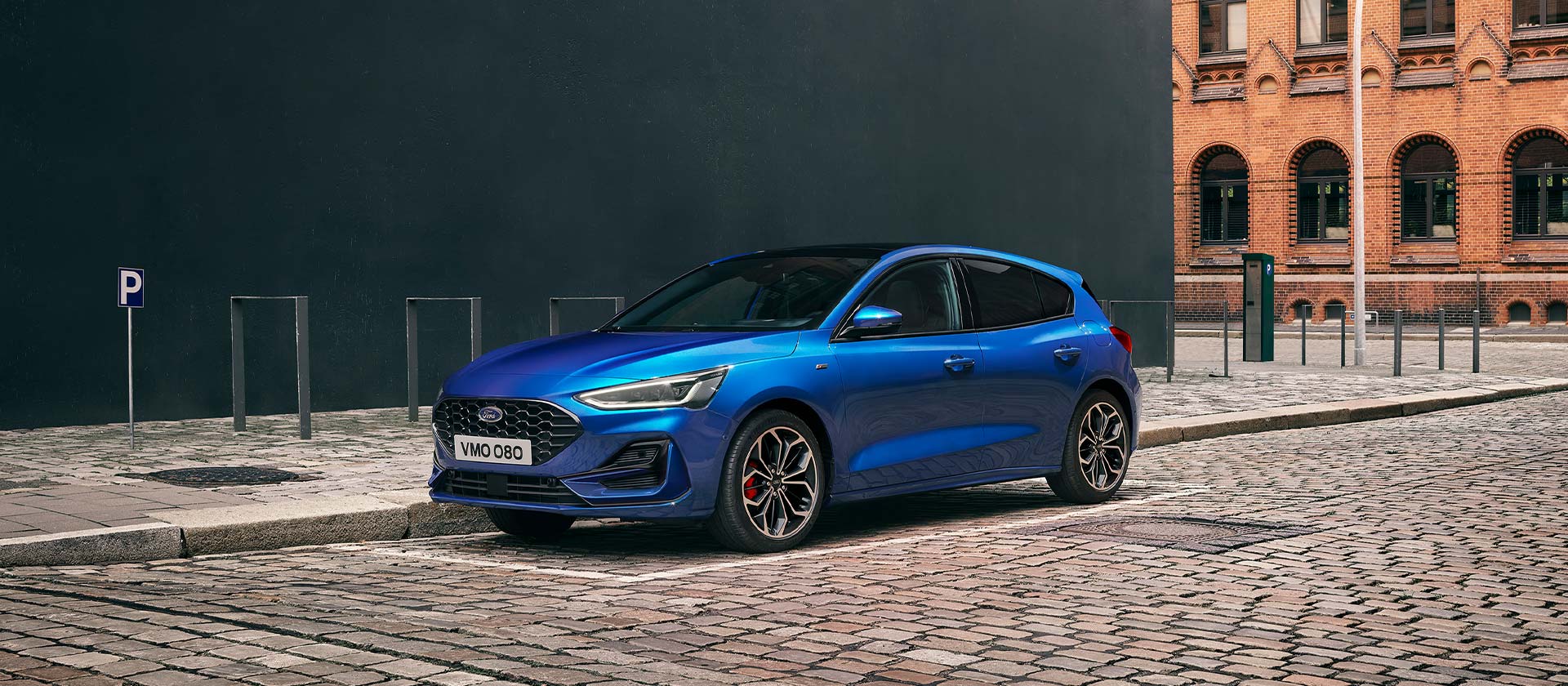 Una Ford Focus modello hatchback blu staziona su una strada acciottolata in ambiente urbano.