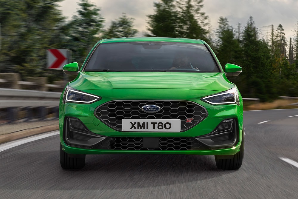 Vista frontale di una Ford Focus ST color Mean Green in curva, con alberi sfocati sullo sfondo.