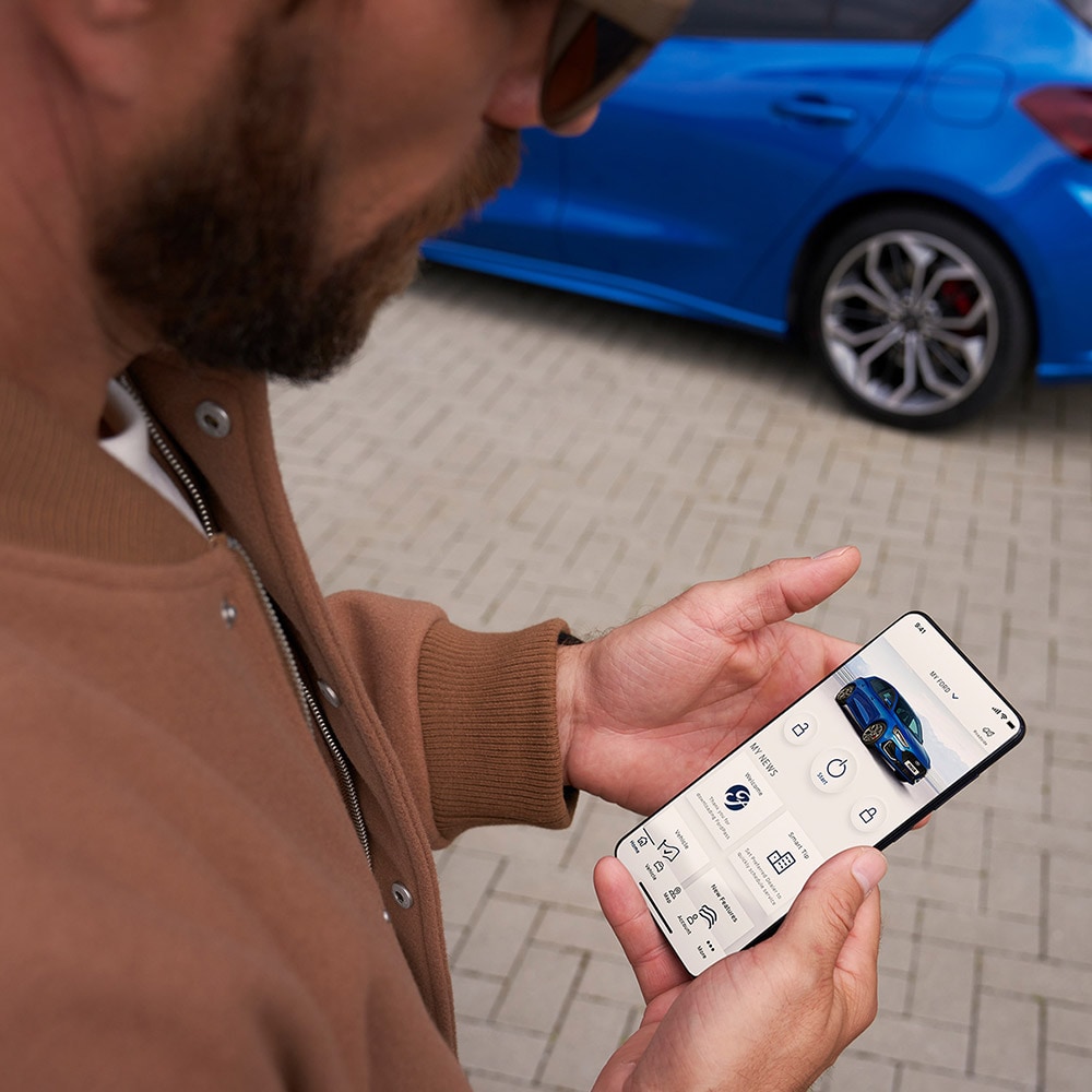 Una persona tiene in mano uno smartphone sul quale è visualizzata l’app FordPass™. Sullo sfondo si trova una Ford Focus blu.