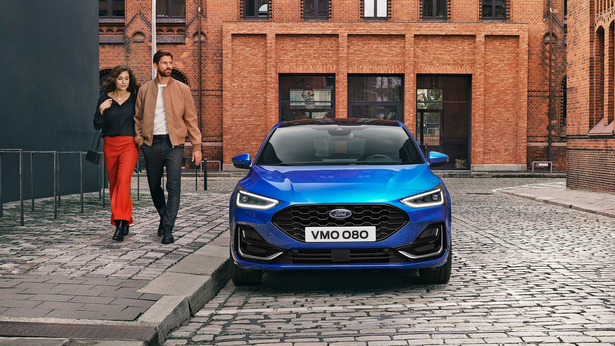 Ford Focus blu. Vista frontale, parcheggiato in un’area industriale. Due persone le passano a fianco.