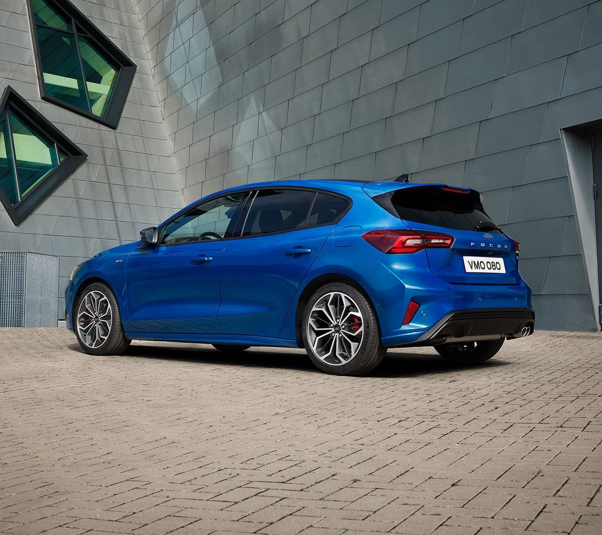 Ford Focus blu. Vista posteriore a tre quarti, parcheggiata davanti a un edificio moderno.