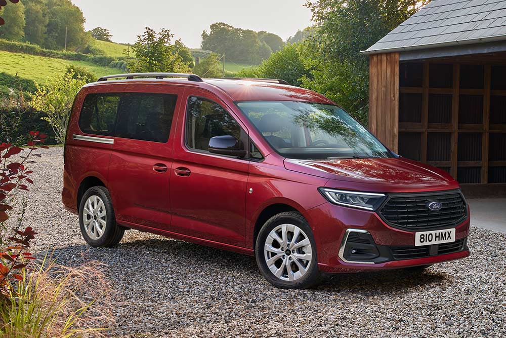 Ford Grand Tourneo Connect rosso, vista frontale a tre quarti, parcheggiato in un vialetto ghiaioso davanti a una casa