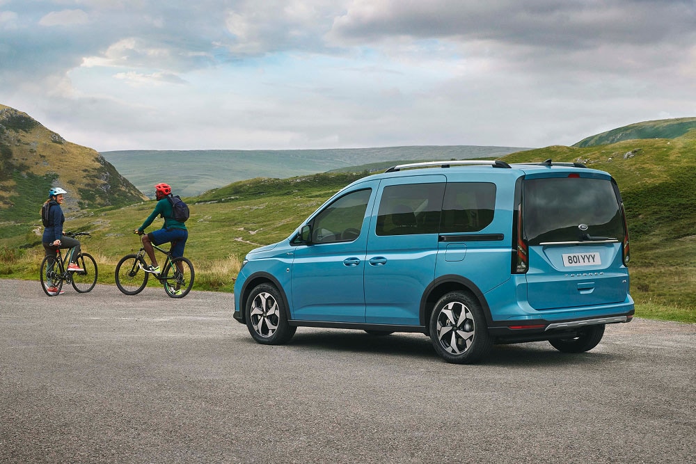 Un Ford Tourneo Connect blu è parcheggiato su una strada in un paesaggio verde e collinare, con due persone in bicicletta nelle vicinanze.