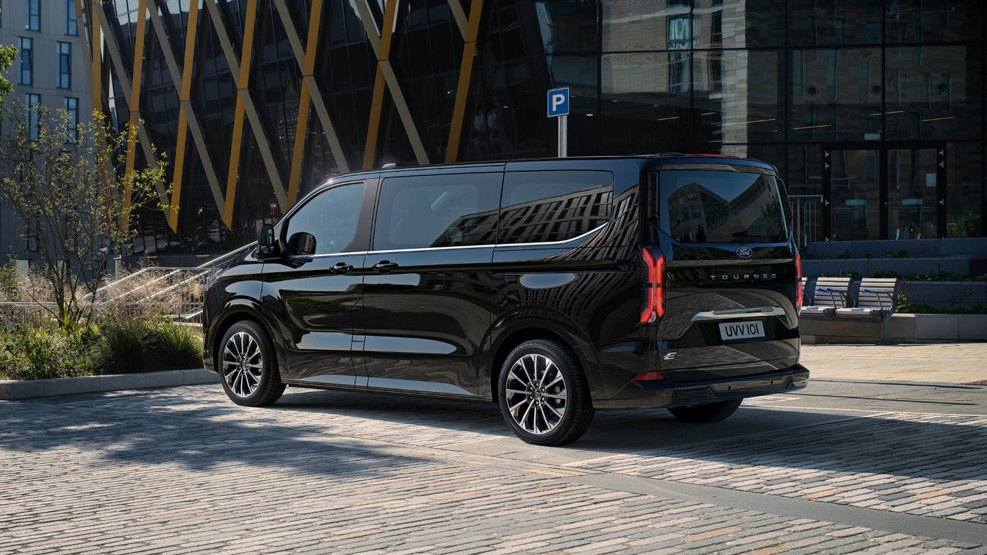 Ford Tourneo Custom nero, vista posteriore a tre quarti, fermo in un parcheggio davanti a edifici moderni