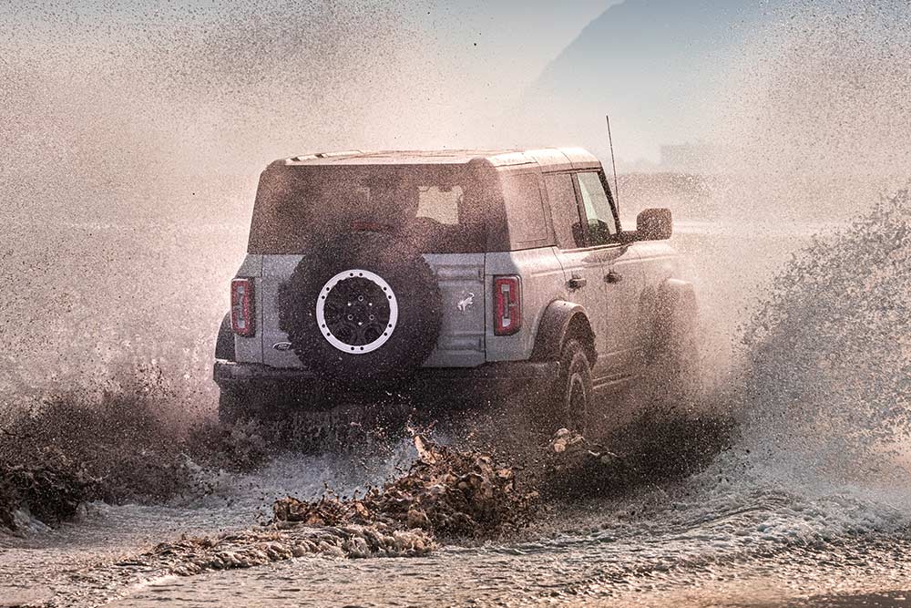 Vista posteriore di un Ford Bronco argento che attraversa spruzzi d'acqua sulla spiaggia.