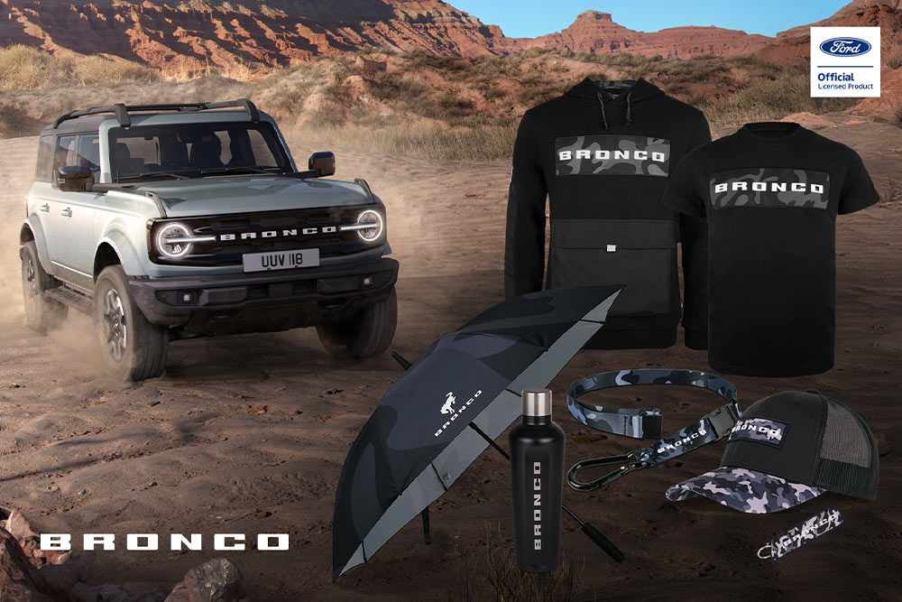Collezione Ford Bronco con t-shirt nera, giacca, polo, cappellino, berretto e borsa impermeabile, con un Bronco grigio sullo sfondo.