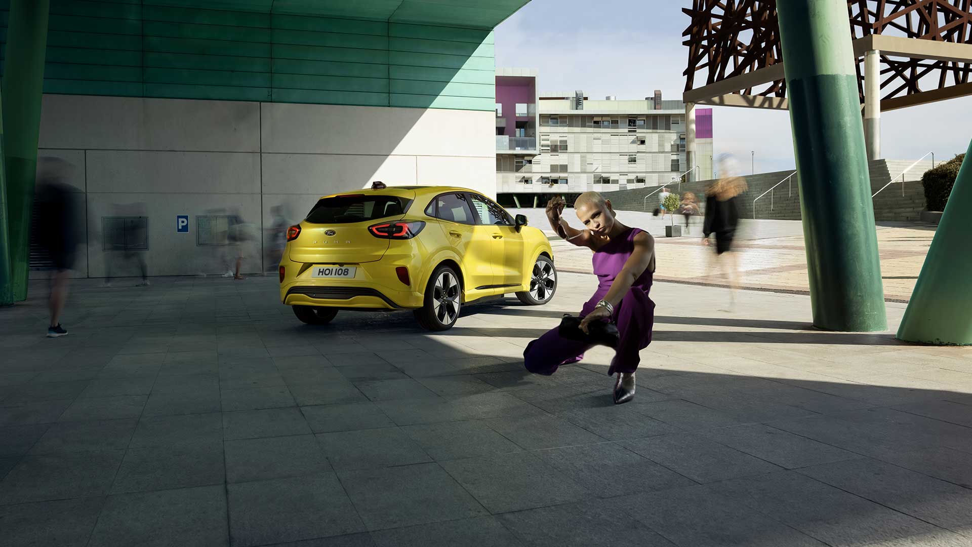 Vista posteriore di una Ford Puma Gen-E® elettrica gialla posteggiata sotto un ponte, accanto a una persona vestita di viola.