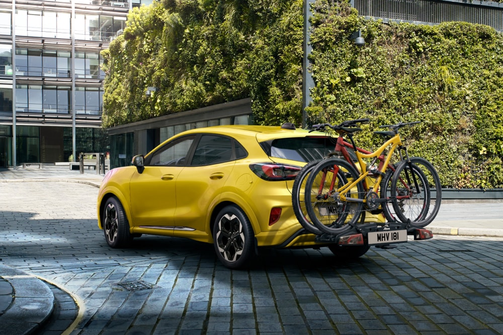 Una Ford Puma Gen-E gialla parcheggiata in città con due biciclette montate su un portabici posteriore.