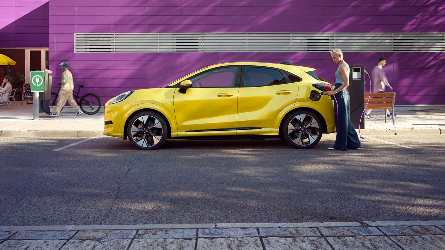 Vista laterale della Ford Puma Gen-E® gialla collegata a una stazione di ricarica pubblica all'interno di un parcheggio.