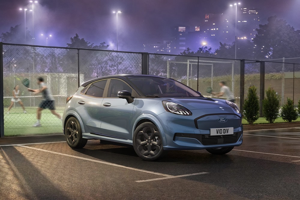 Una Ford Puma Gen-E blu parcheggiata vicino a un campo da tennis con le luci della città sullo sfondo.