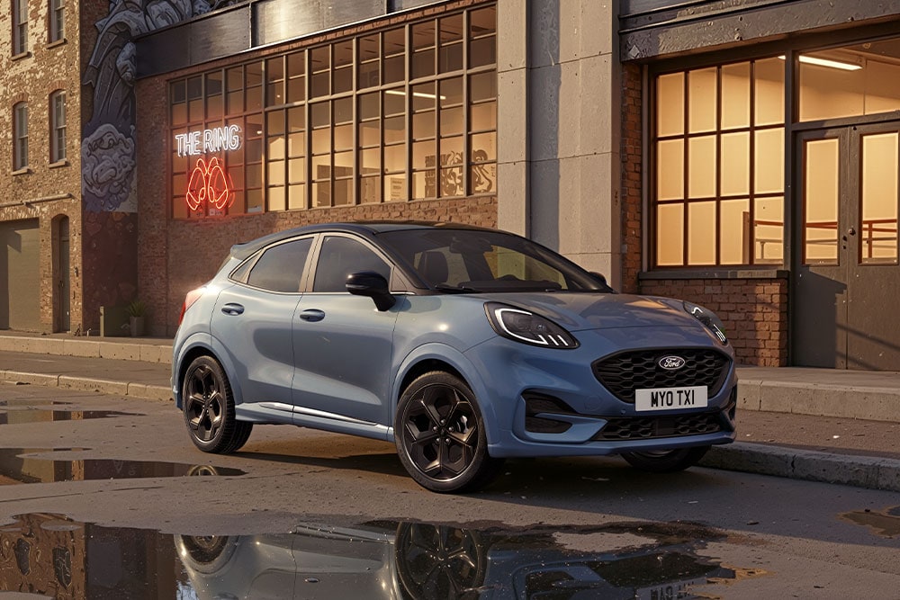 Una Ford Puma blu parcheggiata al tramonto su una strada urbana con pozzanghere nelle quali si riflettono gli edifici.