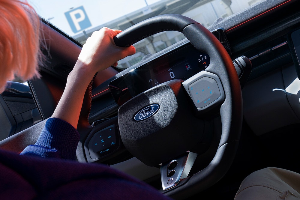 Vista ravvicinata di una donna che impugna il volante di una Ford Capri parcheggiata, di cui è visibile anche il quadro strumenti digitale.