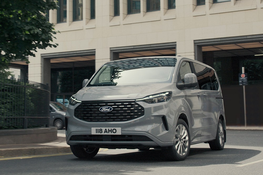 Vista anteriore a tre quarti del Tourneo Custom grigio che guida vicino a un edificio in cemento.
