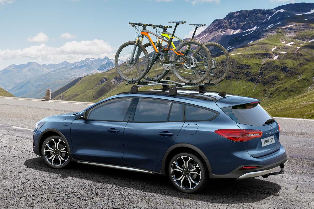 Ford Focus blu, vista posteriore a tre quarti, in marcia su una strada montana con deflettori e tendine parasole.