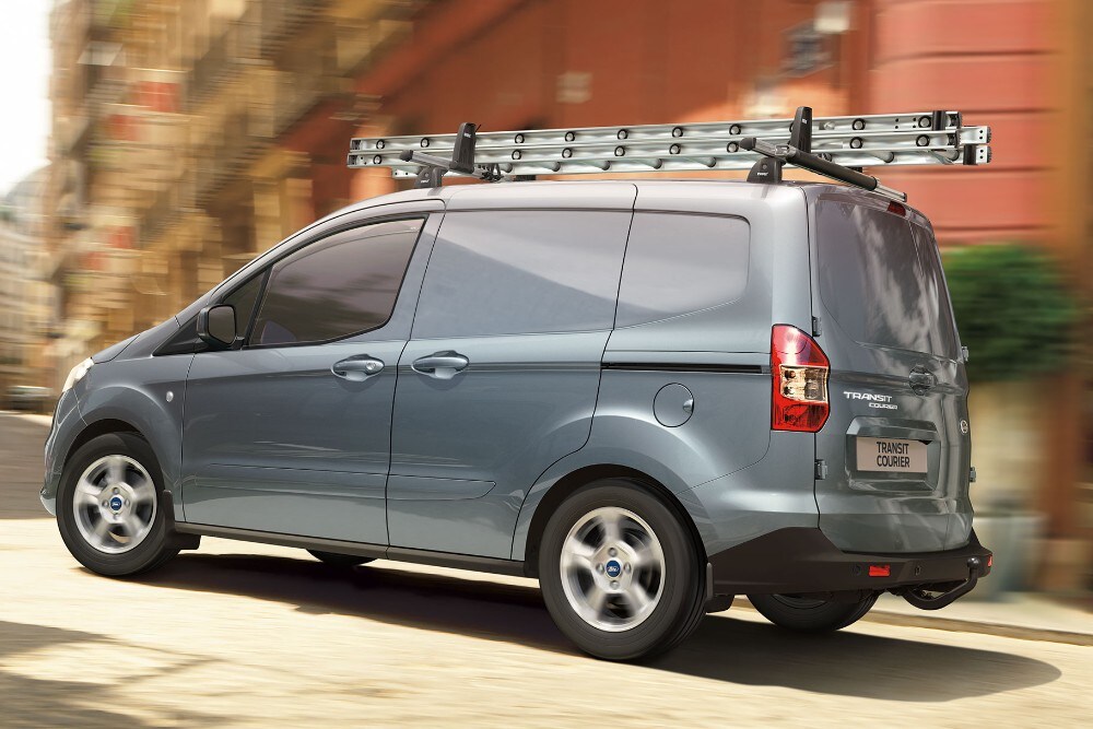 Ford Transit Courier blu argento, vista posteriore a tre quarti, fermo su una strada con barre portapacchi e scala.