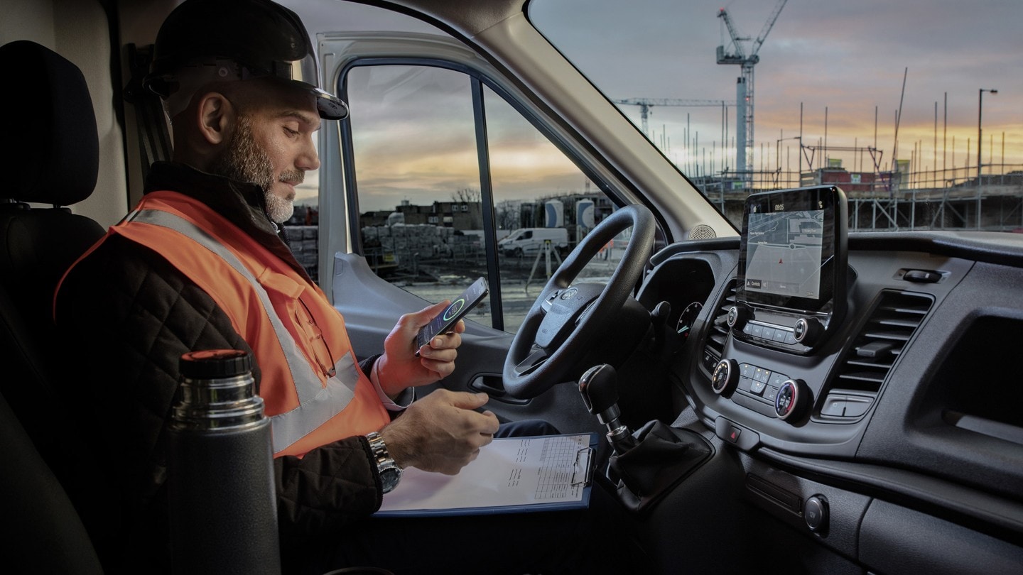 Un operaio con casco da lavoro seduto al volante di un veicolo consulta lo smartphone