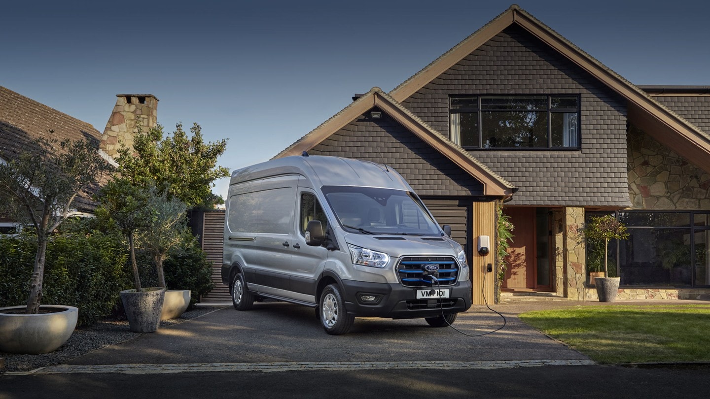 Ford E-Transit color argento, vista frontale a tre quarti, parcheggiato e in carica davanti a un’abitazione