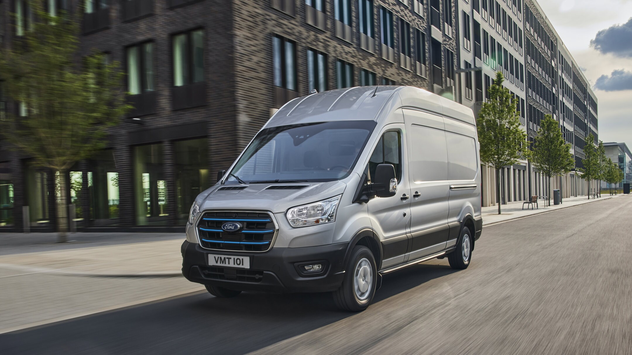 Vista laterale a tre quarti del Ford E-Transit furgone cabinato color argento che costeggia dei grattacieli