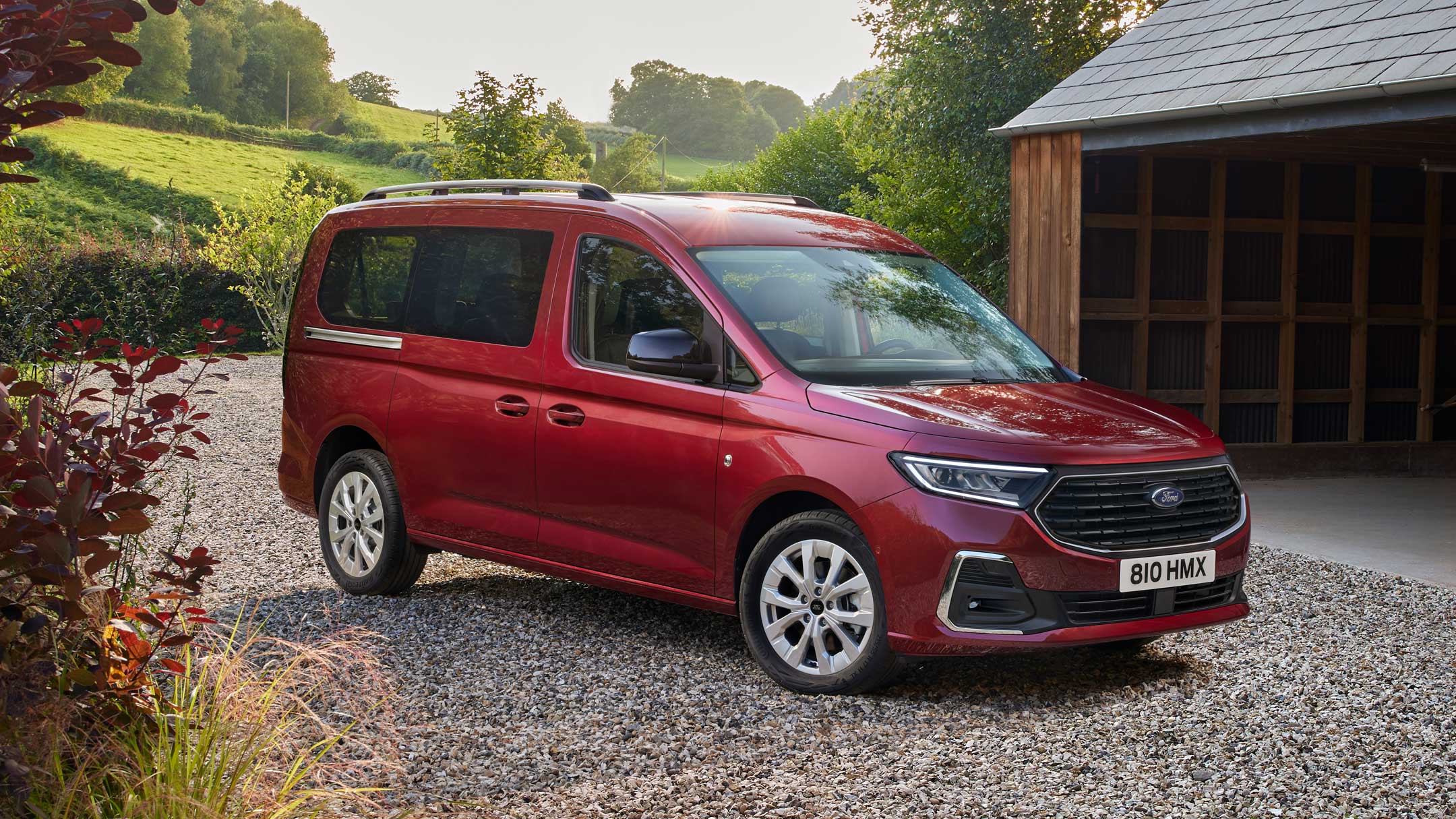 Ford Grand Tourneo Connect rosso, vista frontale a tre quarti, parcheggiato in un vialetto ghiaioso davanti a una casa